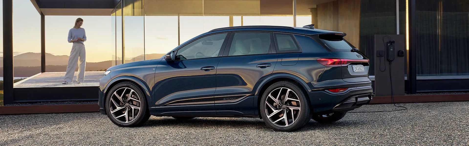 Elektrické Q6 e-tron stojí na štrkovej ploche pred moderným domom so sklenenou fasádou počas západu slnka. Mladá žena v svetlej mikine a bielych nohaviciach stojí na terase domu a pozerá na vozidlo. Na pravej strane je nabíjacia stanica pre elektromobily.