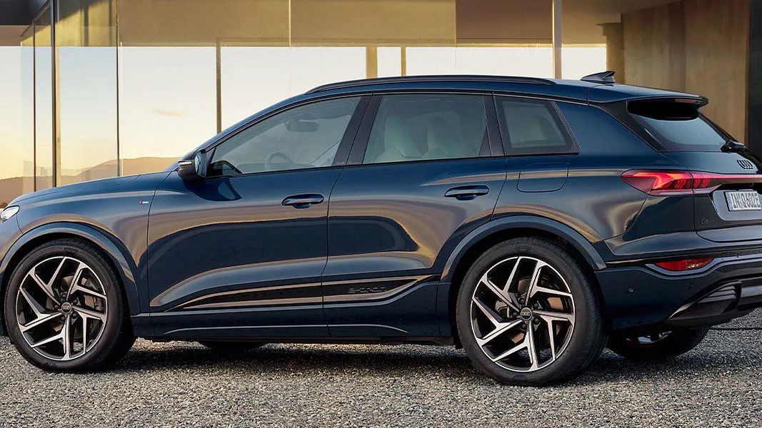 Elektrické Q6 e-tron stojí na štrkovej ploche pred moderným domom so sklenenou fasádou počas západu slnka. Mladá žena v svetlej mikine a bielych nohaviciach stojí na terase domu a pozerá na vozidlo. Na pravej strane je nabíjacia stanica pre elektromobily.
