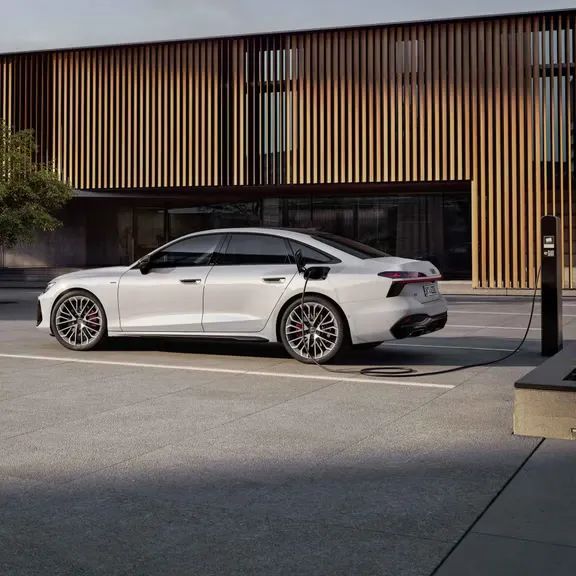 Audi A6 Limuzína e-hybrid zaparkovaná pred modernou budovou na parkovisku a pri verejnej nabíjacej stanici. Vozidlo je pripojené a nabíja sa.