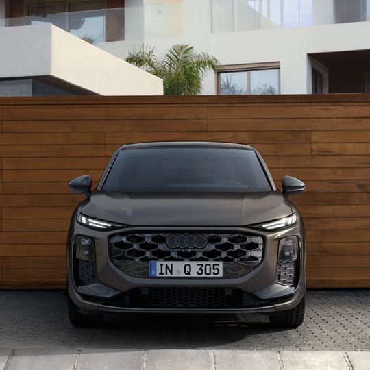 Q3 Sportback z pohľadu spredu zaparkovaný pred modernou budovou, za ním drevený plot..