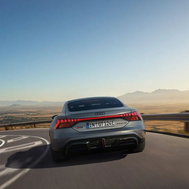 Audi RS e-tron GT počas dynamického prejazdu zákrutou v kopcoch. V pozadí je výhľad na širokú krajinu, malé mesto a vzdialené hory. 