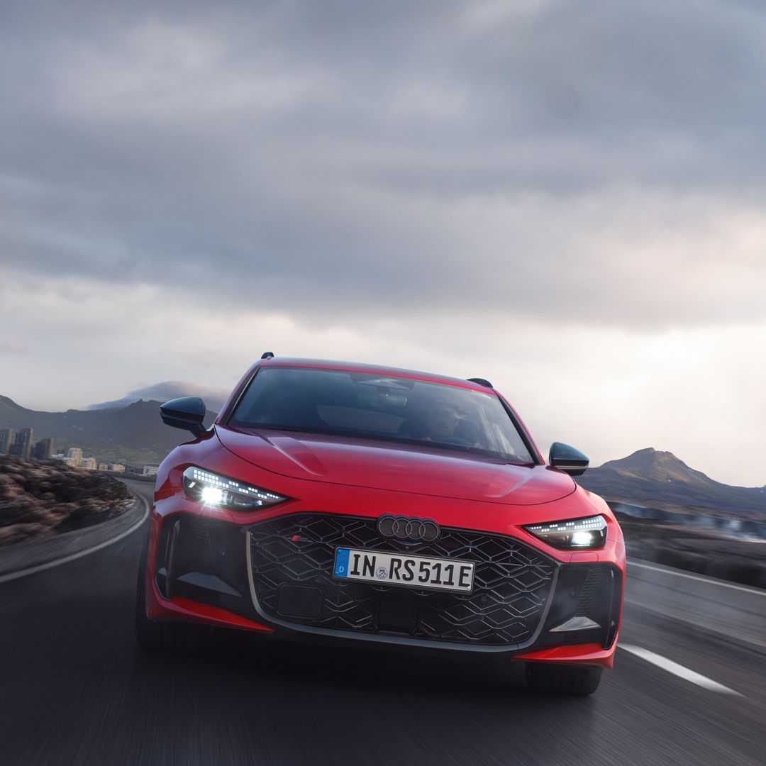 Červené Audi RS 5 Avant jazdí po ceste v horskej krajine. Obloha je zamračená.