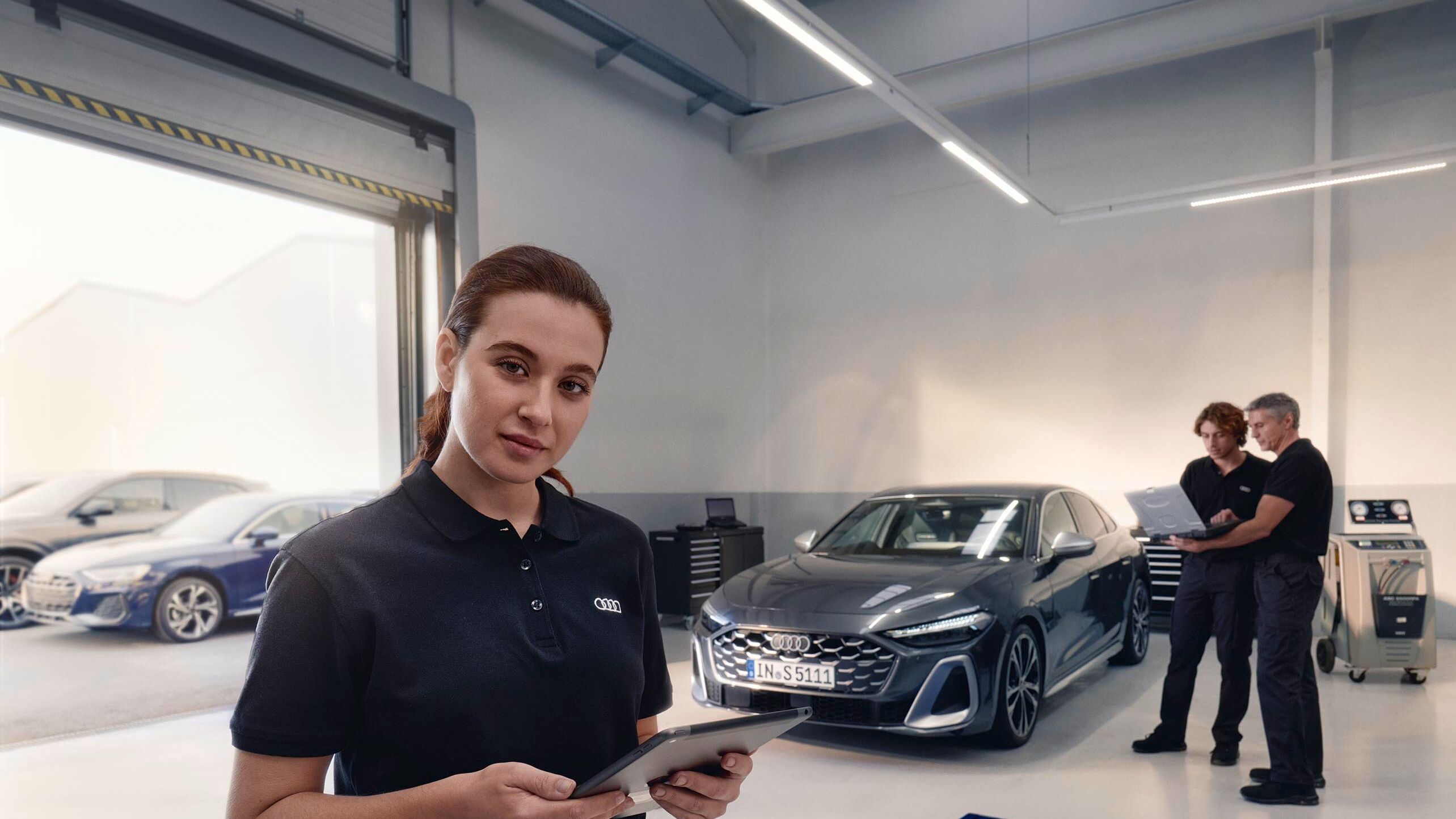 Servisná pracovníčka stojí v servise automobilov a drží tablet. V pozadí sú dvaja muži, ktorí sa rozprávajú pred zaparkovaným autom. Miestnosť je moderná a dobre osvetlená.