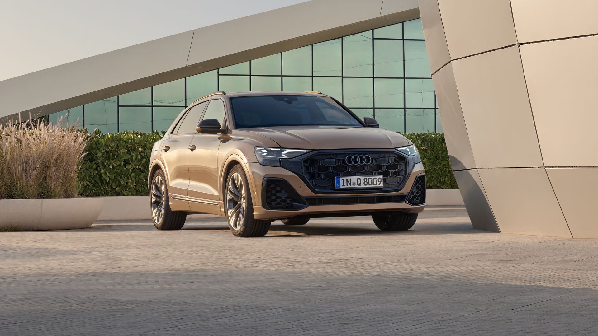 Audi Q8 SUV zaparkované šikmo z prednej strany pred modernou budovou. Vozidlo od budovy za ním, ktorá je celá presklená, oddeľuje živý plot.