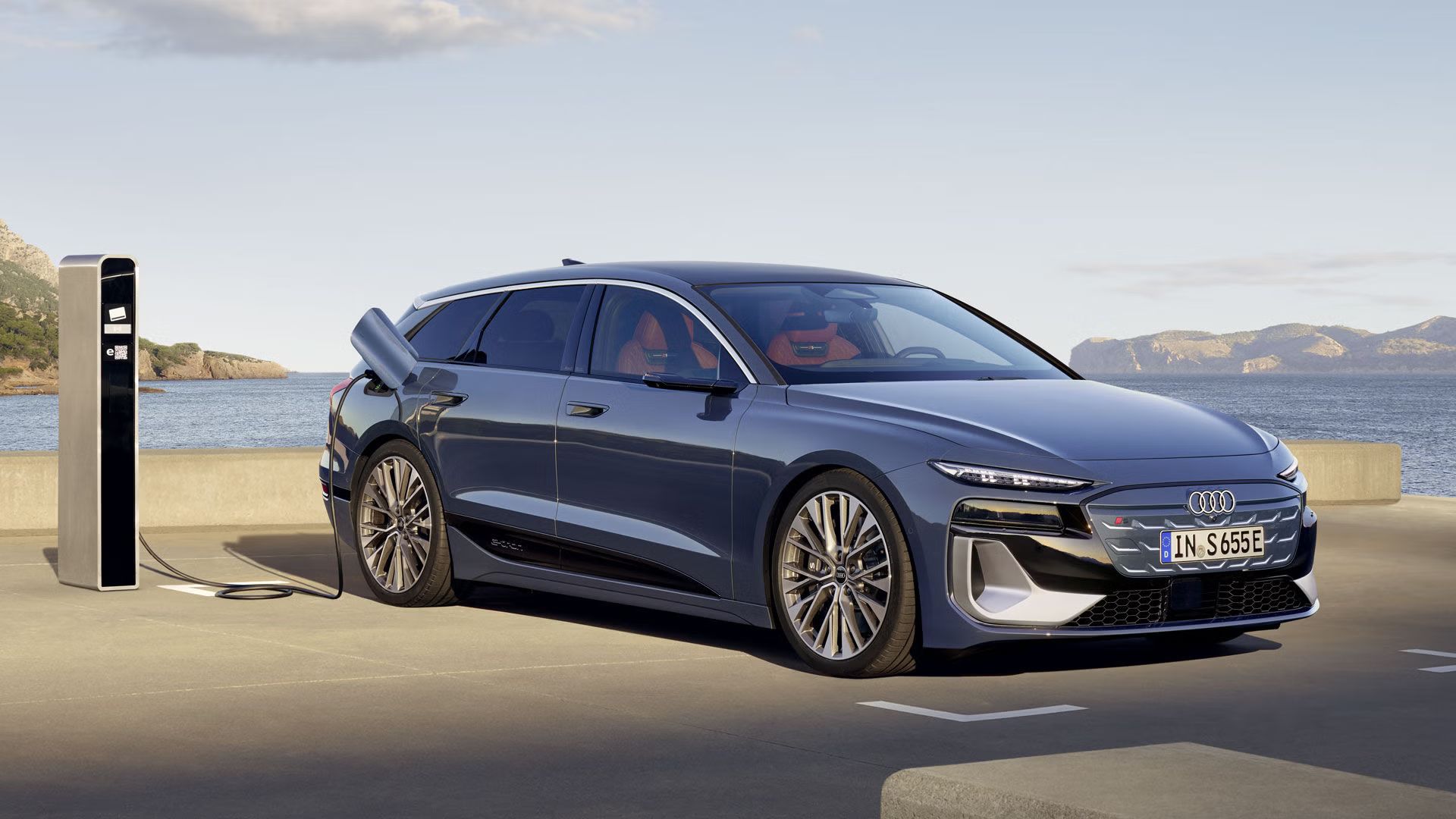 Audi S6 Avant e-tron stojí v zadnej časti parkoviska a je pripojený k verejnej nabíjacej stanici. Za parkoviskom je vidieť more a skaly. 