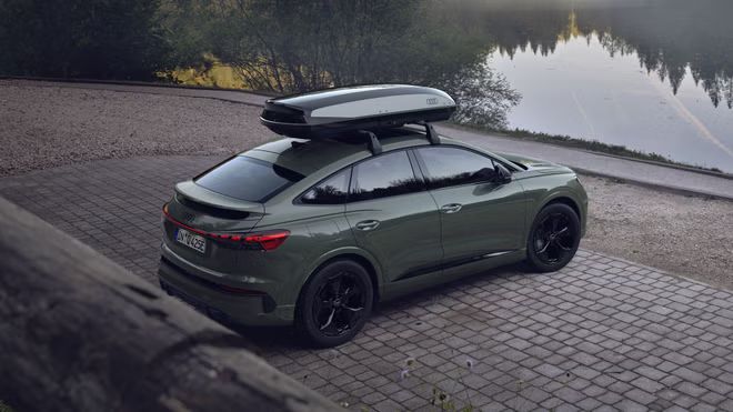 Q4 Sportback e-tron so strešným boxom, snímaný šikmo zozadu na parkovisku pri jazere.