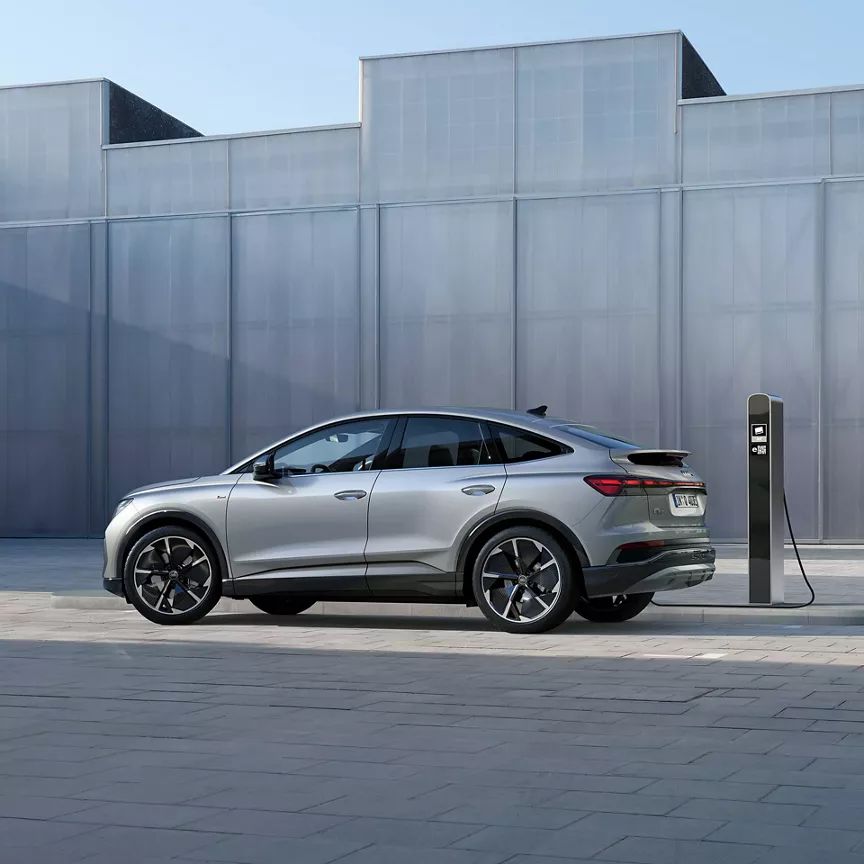 Strieborné Audi Q4 je pripojené k nabíjacej stanici a stojí na modernom dláždenom námestí. V pozadí sú viditeľné veľké sivé budovy. Scéna pôsobí pokojne a futuristicky.