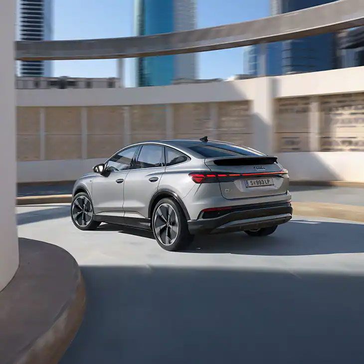 Strieborné elektrické SUV Audi Q4 jazdí po parkovacom dome. V pozadí sú viditeľné vysoké budovy. Auto má elegantný dizajn s aerodynamickými líniami.