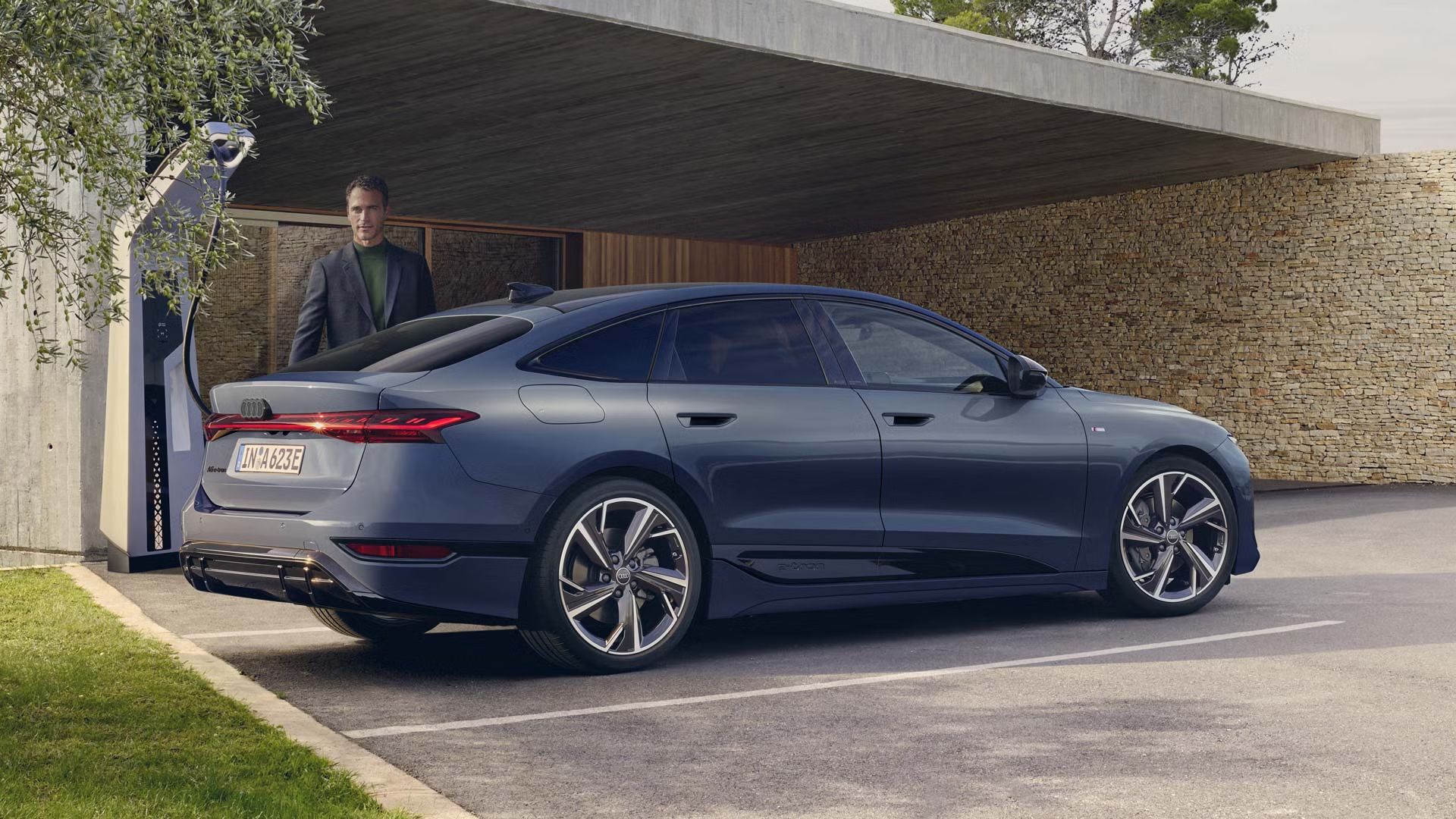 A6 Sportback e-tron stojí v zadnej časti parkoviska pri nabíjacej stanici. V pozadí je vidieť modernú budovu a muža v obleku.