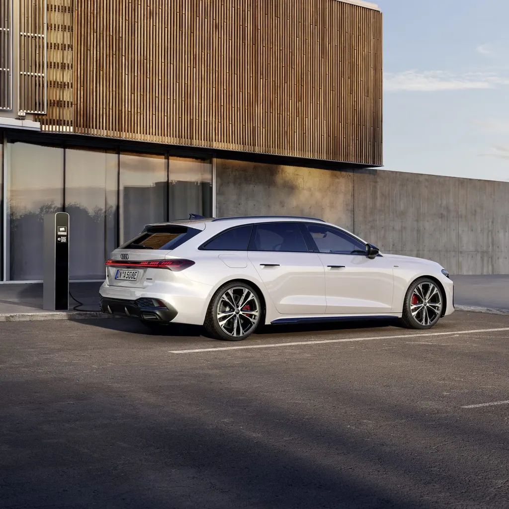 Audi A5 Avant e-hybrid zaparkovaný pri chodníku vedľa budovy s drevenou, sklenenou a betónovou fasádou. Vozidlo je pripojené k nabíjacej stanici a nabíja sa.