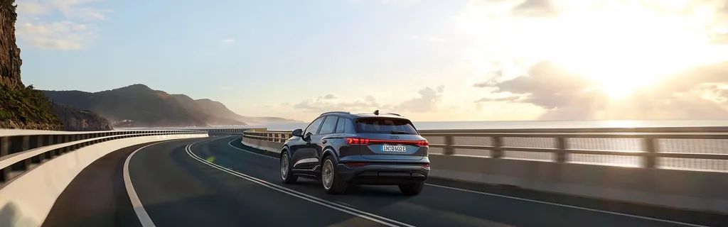 Audi Q6 SUV e-tron počas jazdy po moste. V pozadí je pohľad na pobrežie a more.
