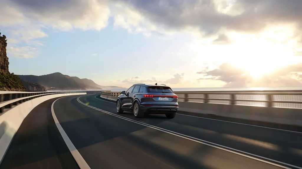 Audi Q6 SUV e-tron počas jazdy po moste. V pozadí je pohľad na pobrežie a more.