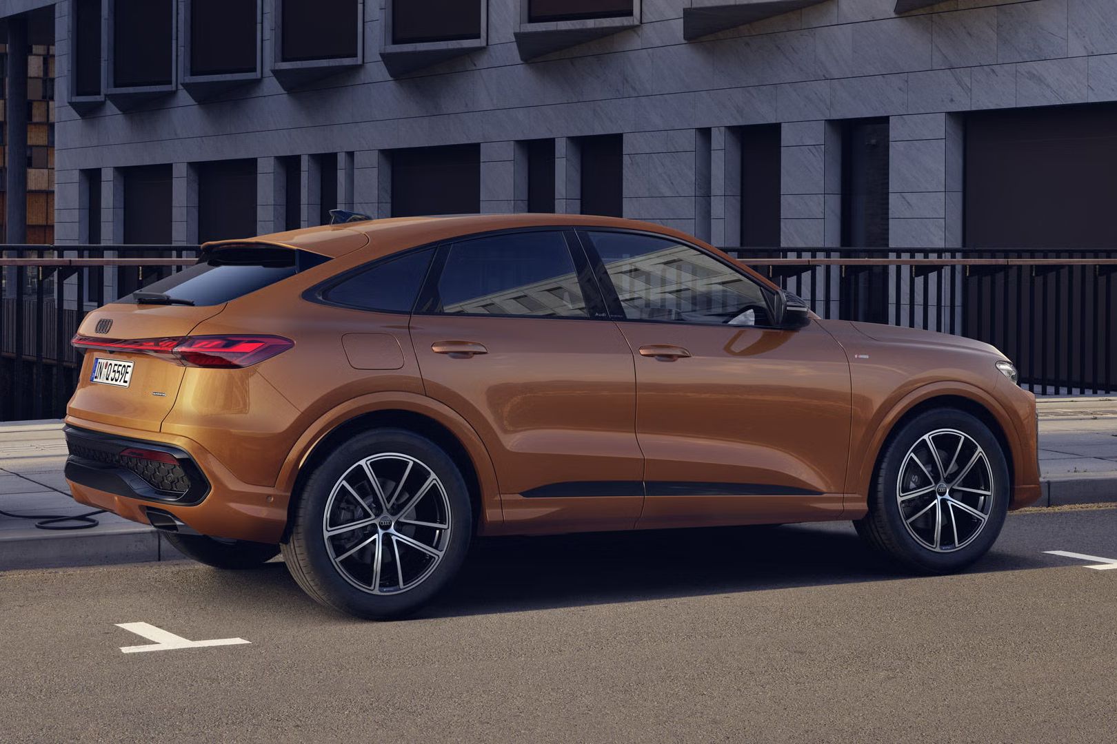 Audi Q5 Sportback e-hybrid s individuálnym lakovaním v hnedom metalízovom odtieni Ipanema stojí pri ceste a je pripojené k nabíjacej stanici.