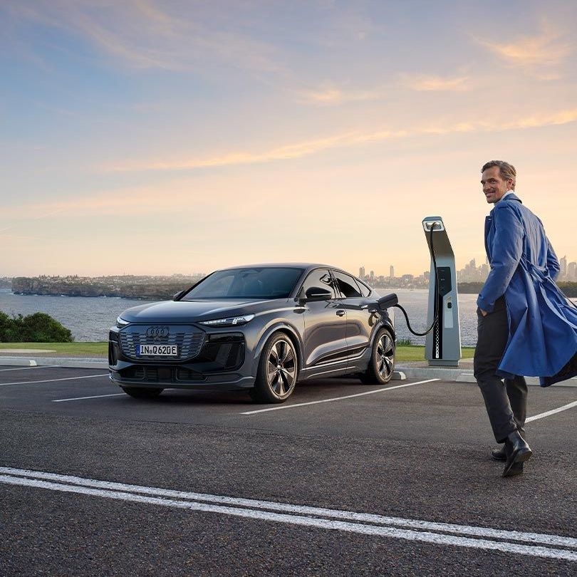Muž kráča k sivému Audi Q6 Sportback e-tron, ktoré stojí pri nabíjacej stanici.