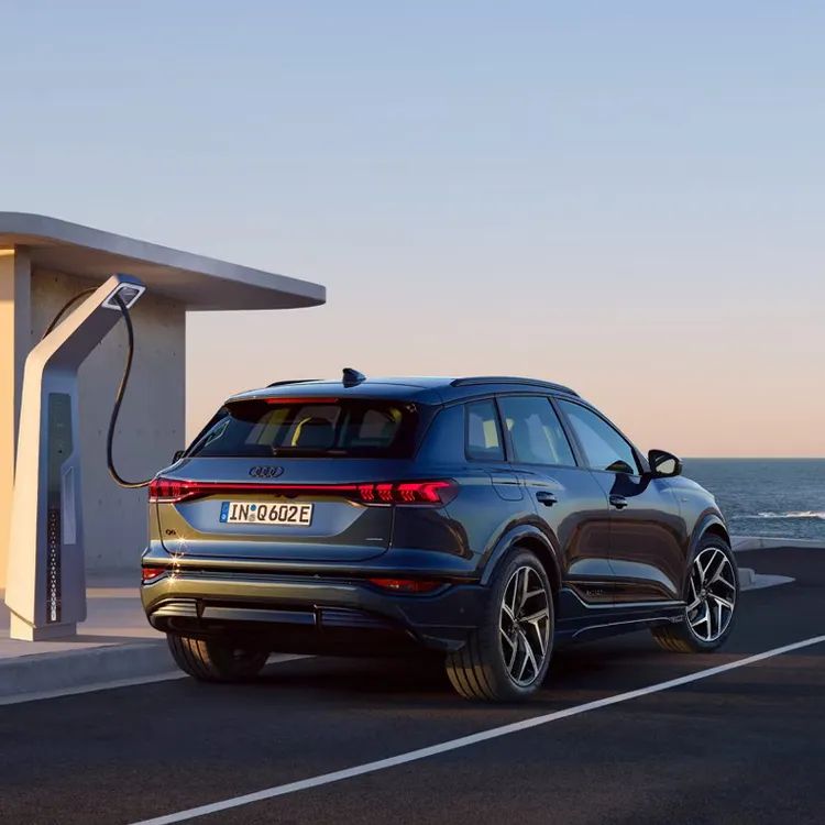 Športové Audi Q6 SUV je zaparkované pri nabíjacej stanici na parkovisku s výhľadom na more. Je napájaný elektrickým káblom. Na pozadí je jasná obloha pri západe slnka.