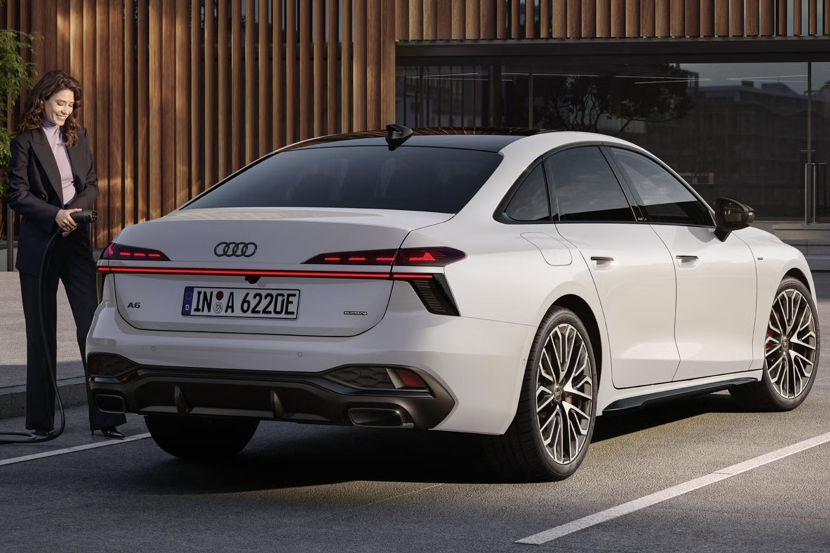 Biele Audi A6 Limuzína e-hybrid stojí na parkovisku pred budovou. Mladá žena pripája limuzínu A6 Limuzína e-hybrid k nabíjacej stanici.