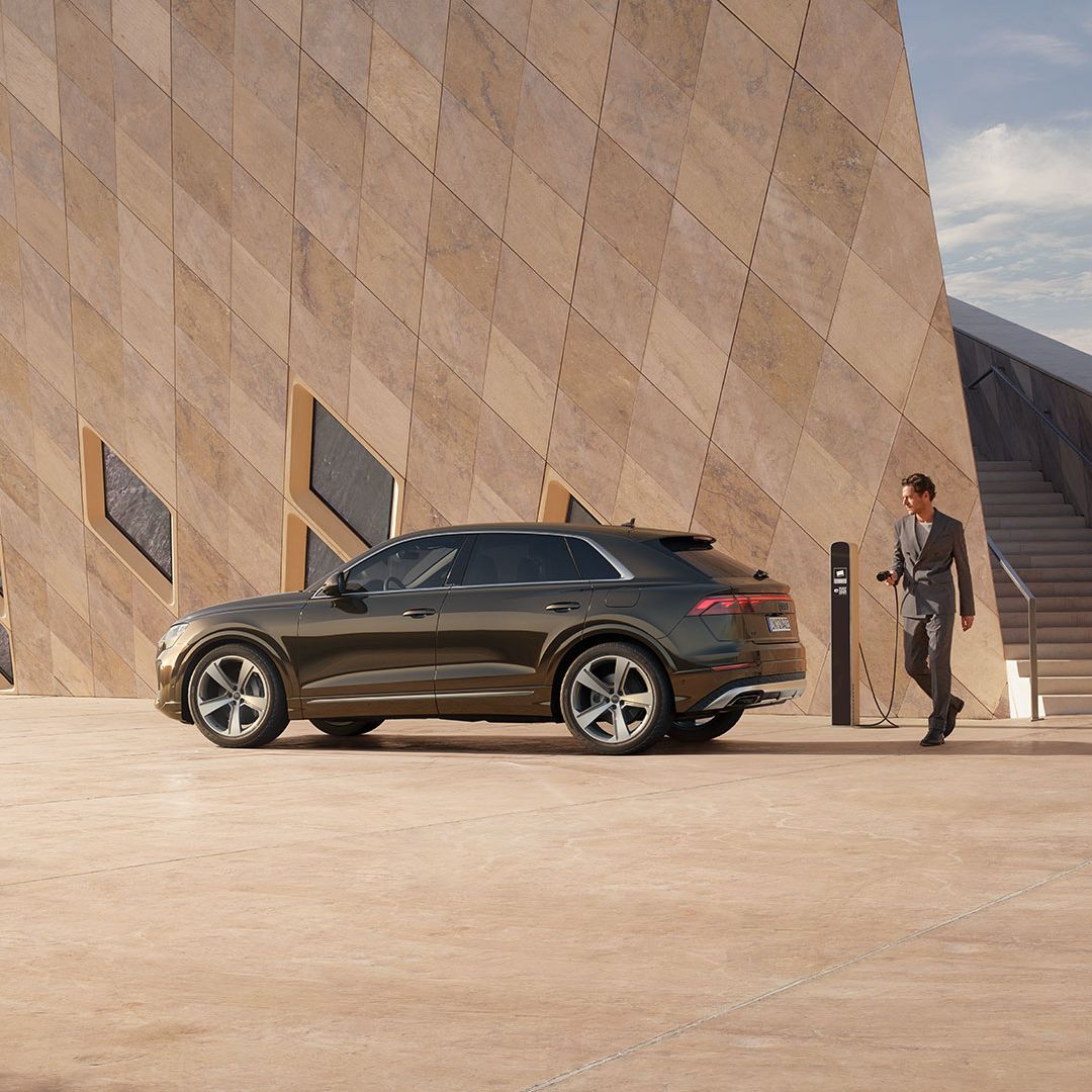 Audi Q8 SUV TFSI e je zaparkovaný pri modernej verejnej budove. Vedľa budovy stojí verejná nabíjacia stanica, muž drží v ruke konektor a ide vozidlo pripojiť k nabíjacej stanici.