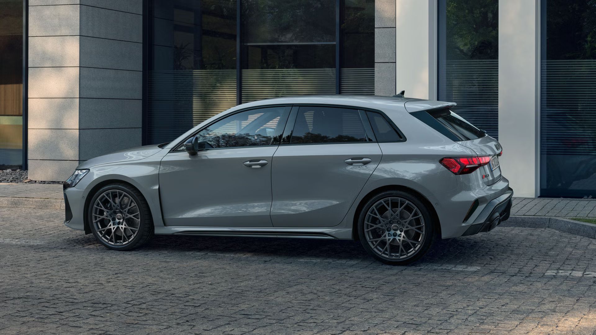 RS 3 Sportback s individuálnym lakovaním v šedom odtieni Pfeil s perleťovým efektom, zaparkovaný zboku na dlažobných kockách, v pozadí obrysy modernej budovy.