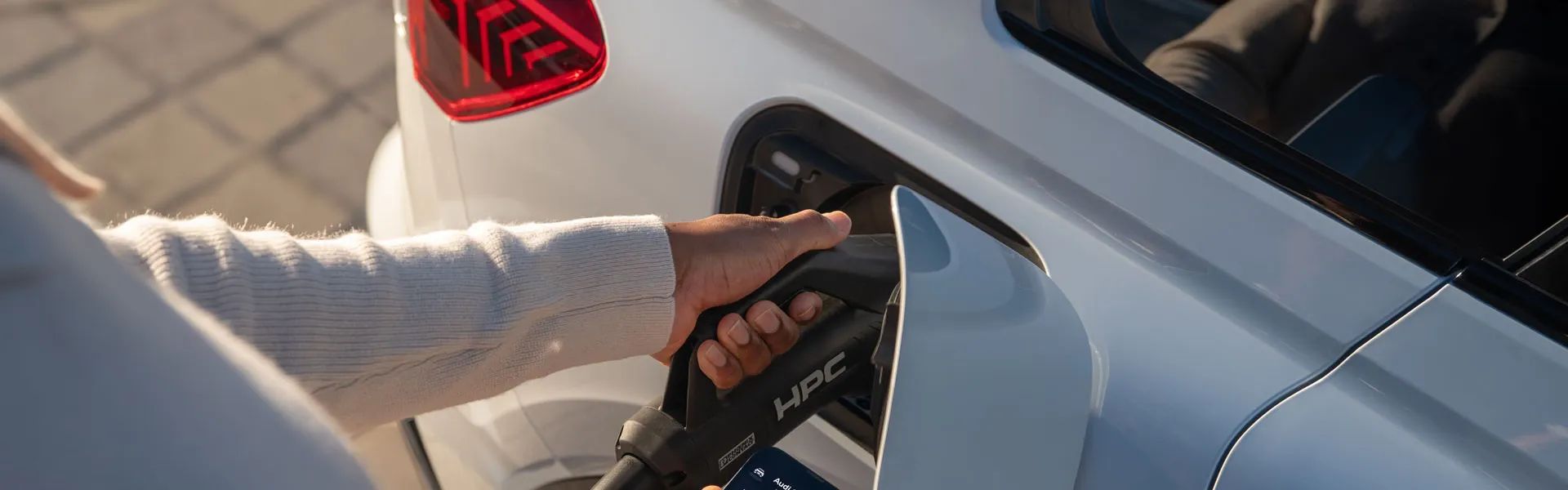 Ruka drží nabíjací konektor a pripája ho do elektrického vozidla. Na pozadí vidieť zadné svetlá auta. Nabíjací konektor má označenie "HPC".