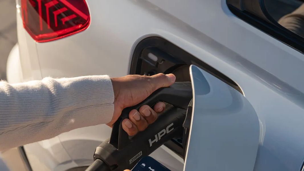 Ruka drží nabíjací konektor a pripája ho do elektrického vozidla. Na pozadí vidieť zadné svetlá auta. Nabíjací konektor má označenie "HPC".