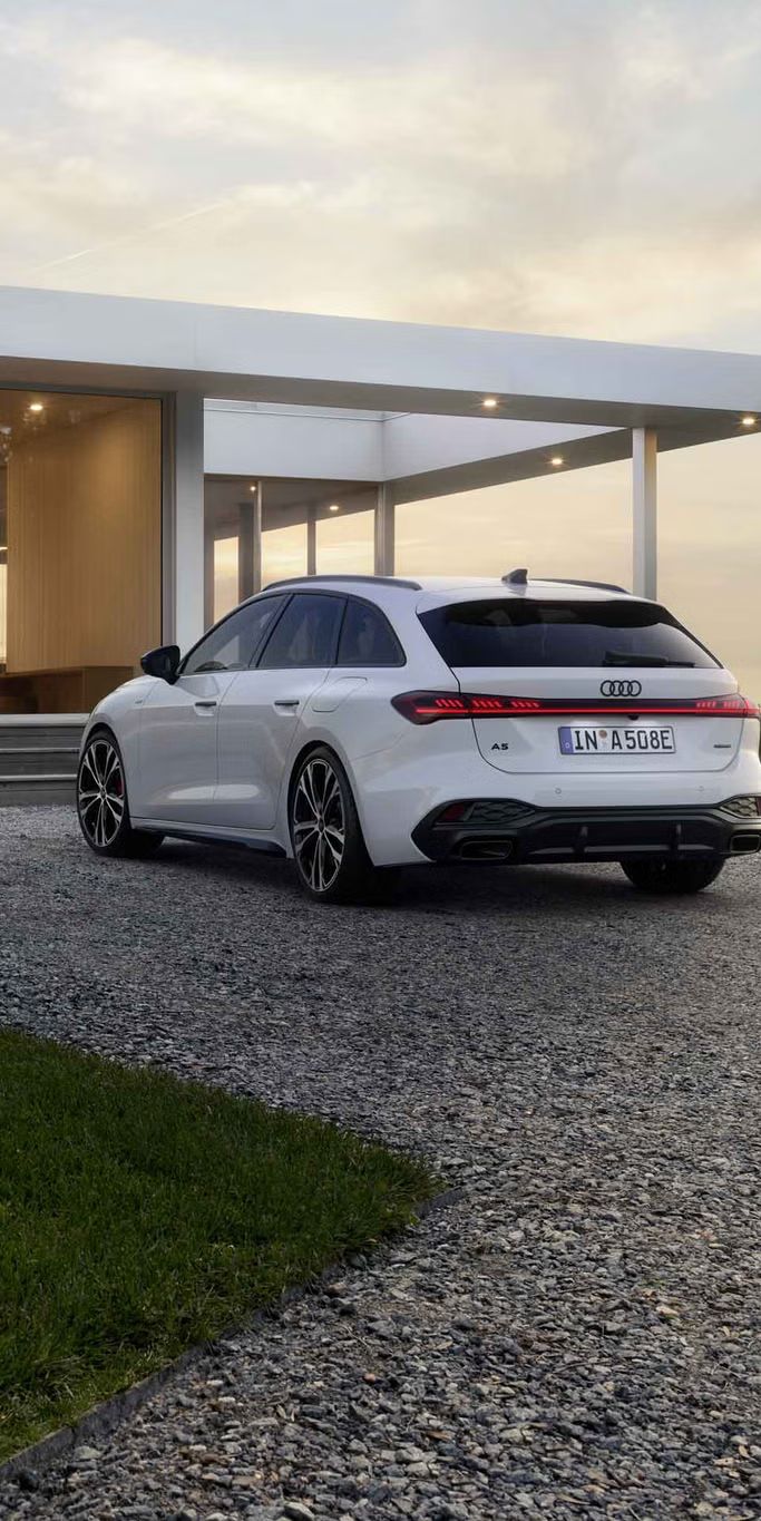 Der A5 Avant e-hybrid steht in einer schrägen Heckperspektive auf einer Kiesfläche vor einem modernen Wohnhaus.