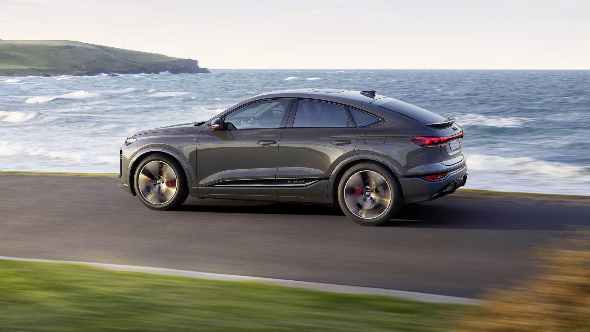 Bočný pohľad na model Q6 Sportback e-tron, ktorý jazdí po ceste popri mori.
