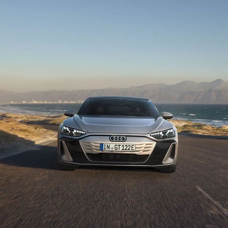 Audi S e-tron GT na ceste popri pobreží. V pozadí vidieť vyššie kopce, vzdialené mesto na pobreží a rozbúrenú hladinu mora.