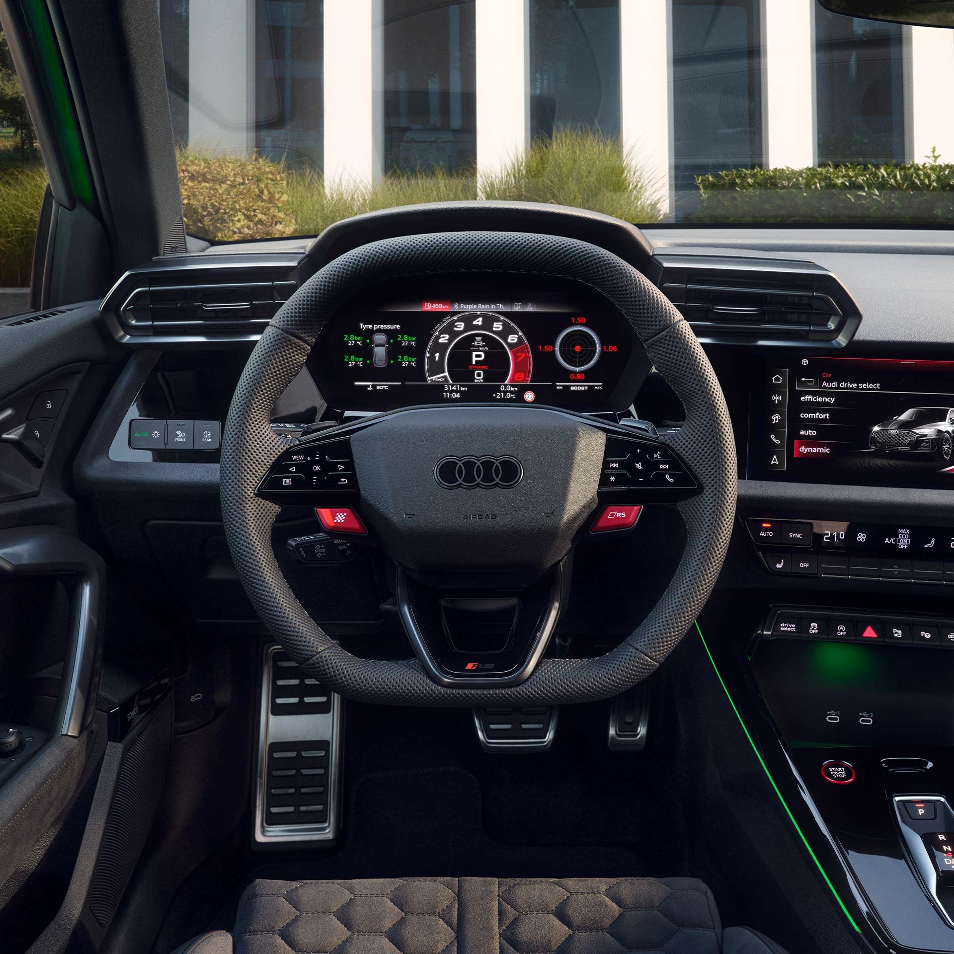 Pohľad na športový, multifunkčný volant, digitálny prístrojový panel, displej MMI a stredovú konzolu. Cez čelné sklo je výhľad na verejnú budovu.