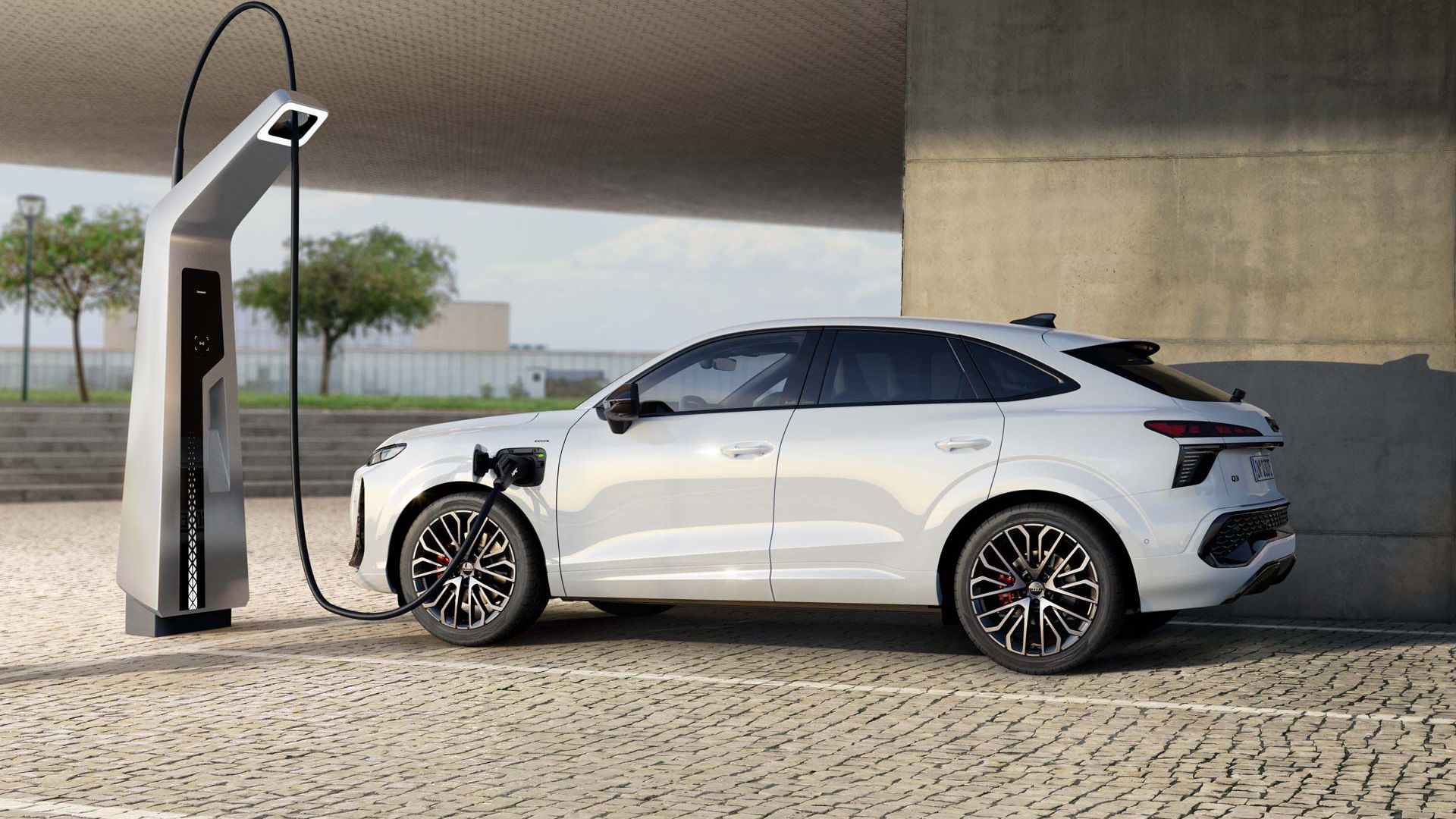 Biele Audi Q3 Sportback e-hybrid sa nabíja na nabíjacej stanici. Vozidlo stojí na dláždenom parkovisku pod veľkou budovou. V pozadí sú vidieť stromy a zamračenú oblohu.