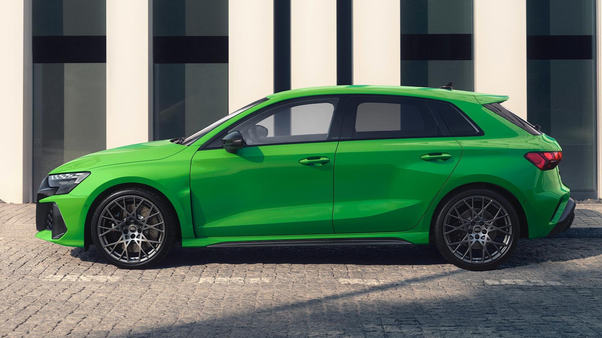 Zelené Audi RS 3 Sportback zaparkované na dlažbe pri chodníku vedľa modernej, verejnej budovy.