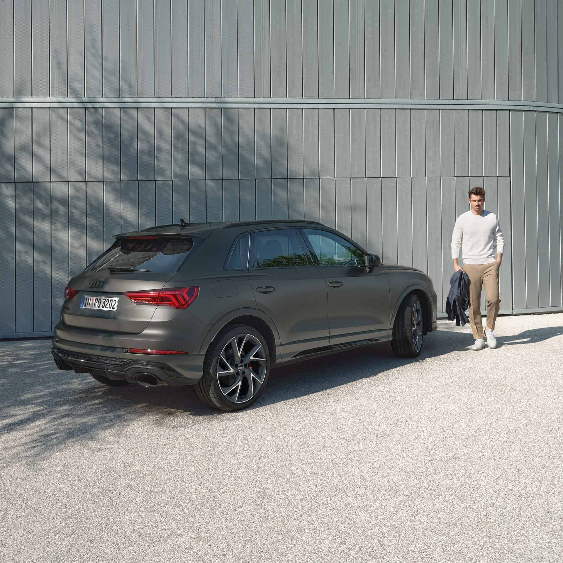 Šedé RS Q3 zaparkované pred modernou stavbou a muž prechádza popri vozidlu s bundou v ruke. Automobil stojí v tieni veľkého stromu.