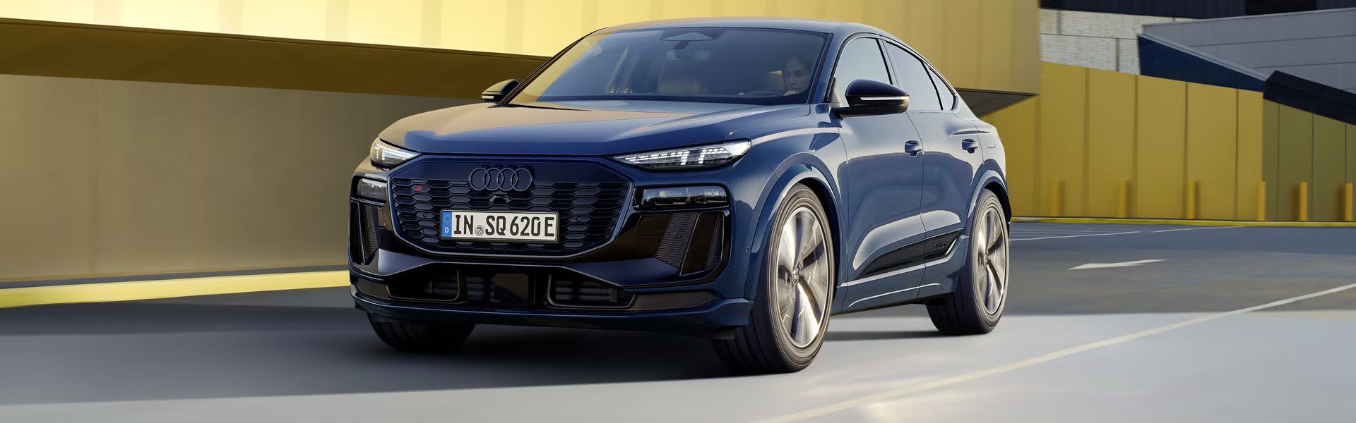 Audi SQ6 Sportback e-tron počas jazdy na ceste v meste. Za vozidlom je vidieť budovy. V popredí je jedna so žltou fasádou, za ňou ďalšia s čiernou fassádou.