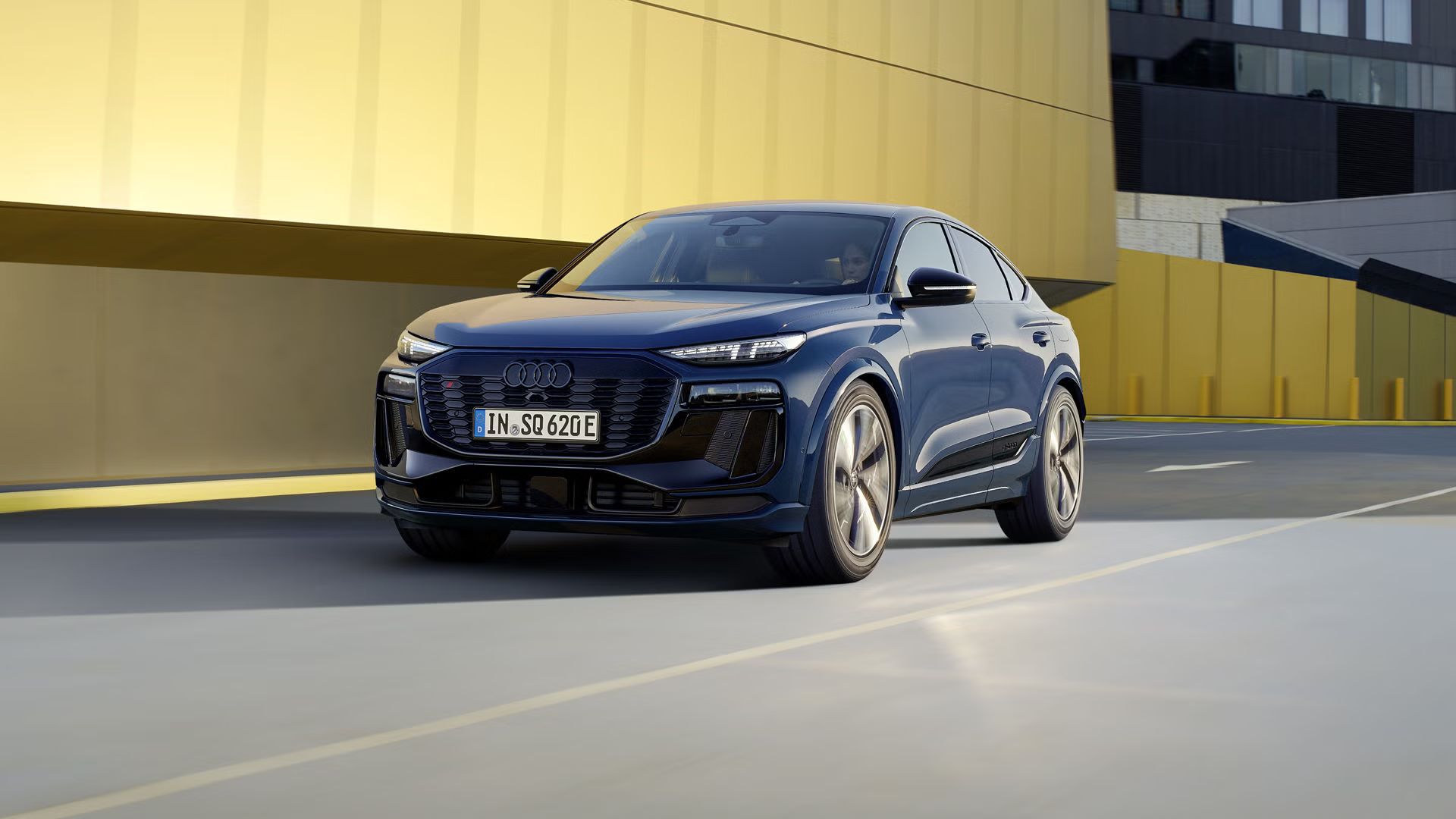 Audi SQ6 Sportback e-tron počas jazdy na ceste v meste. Za vozidlom je vidieť budovy. V popredí je jedna so žltou fasádou, za ňou ďalšia s čiernou fassádou.