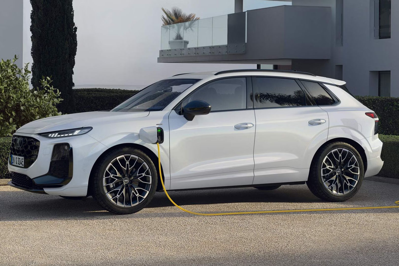 Bočný profil bieleho Audi Q3 SUV e-hybrid, ktorý sa nabíja na parkovisku pred moderným domom.