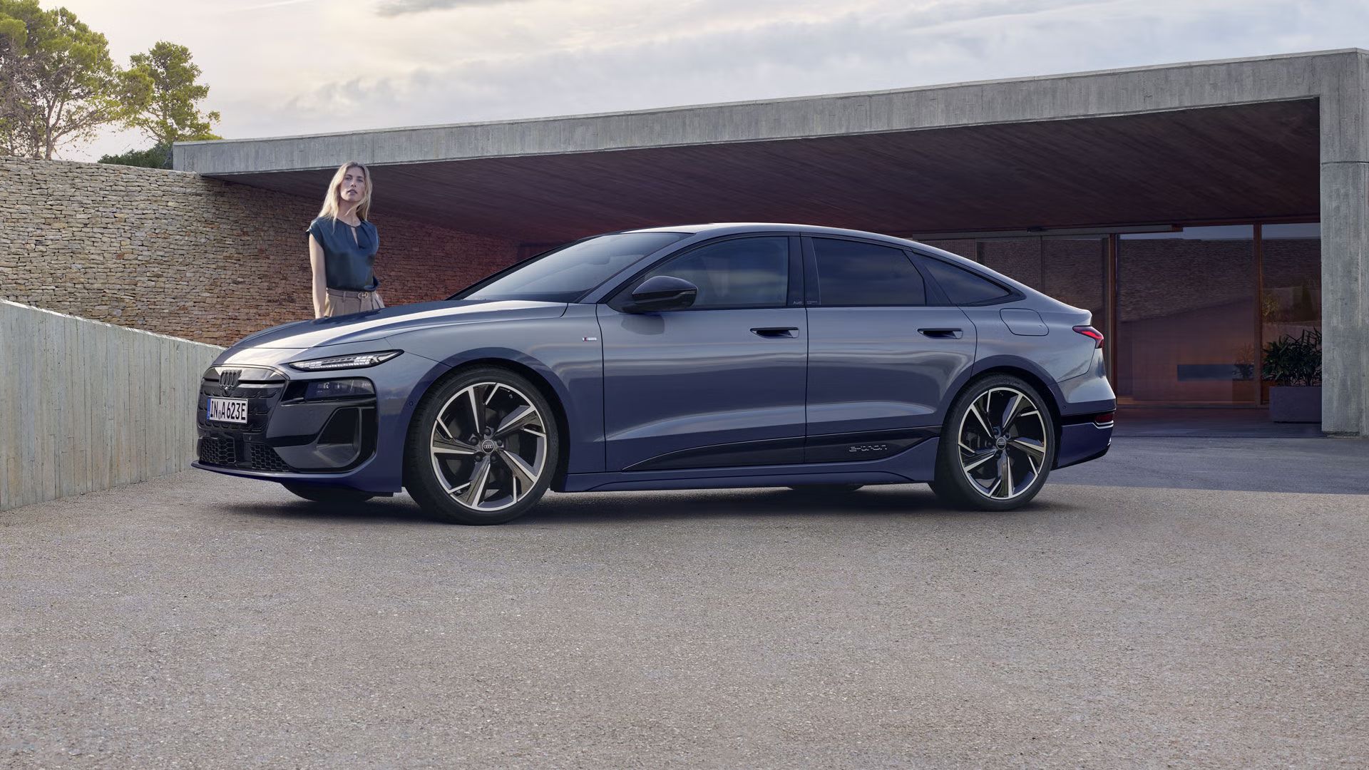 Audi A6 Sportback e-tron z bočného pohľadu je zaparkovaný pred modernou, betónovou budovou s jednou kamennou stenou. 