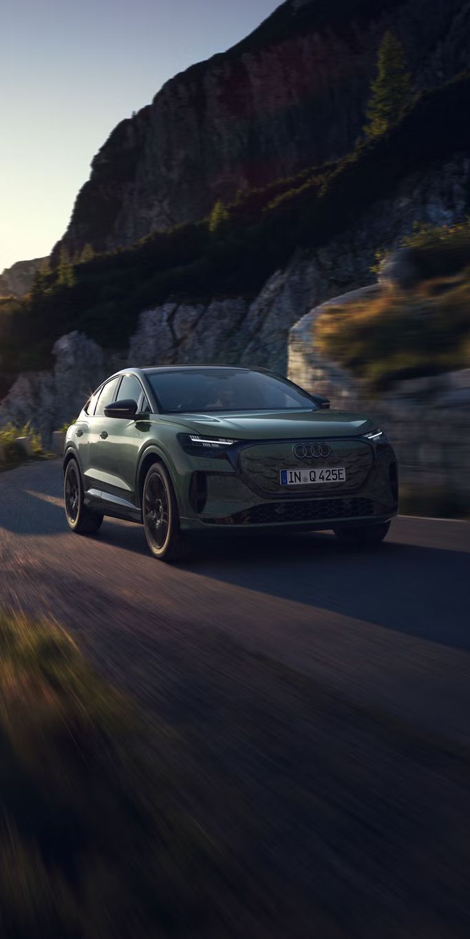 Q4 Sportback e-tron počas jazdy po horskej ceste. Slnečné lúče zľava hore osvetľujú obraz, vozidlo vrhá tieň dopredu.
