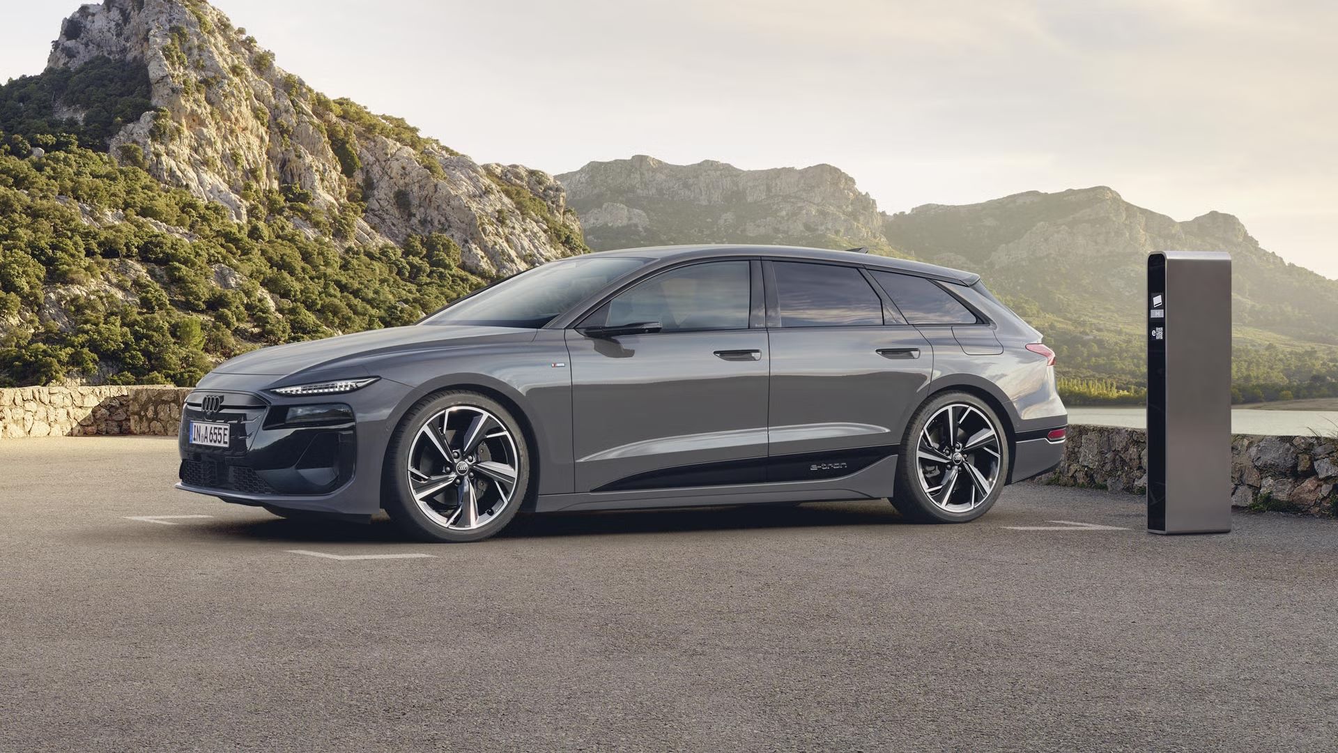 A6 Avant e-tron stojí na štrkovom parkovisku pred jazerom a je pripojený k nabíjacej stanici. V pozadí sú vidieť hory porastené stromami.