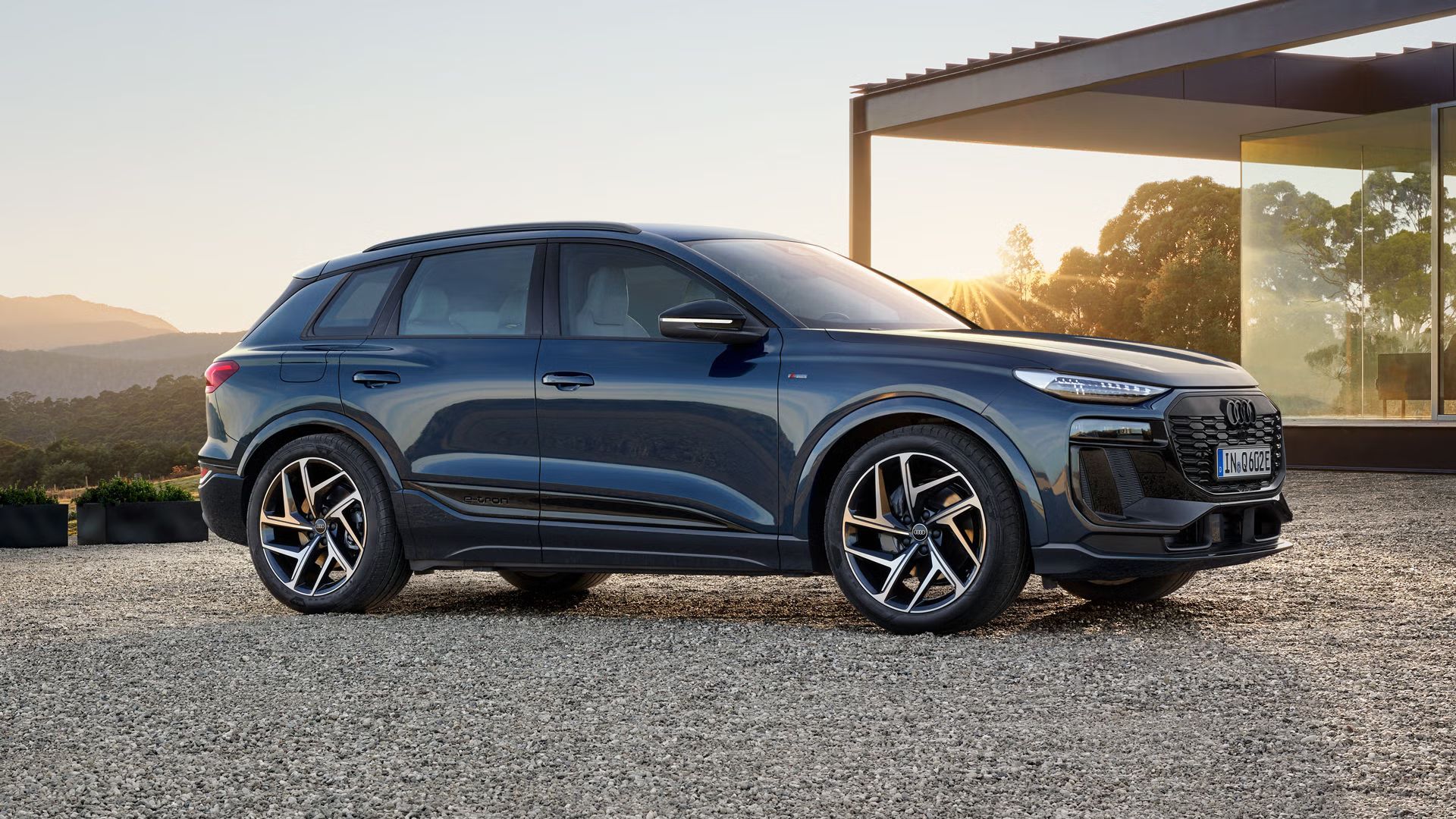 Audi Q6 e-tron SUV stojí bokom na štrkovom priestranstve pred moderným domom a osvetľuje ho zapadajúce slnko.