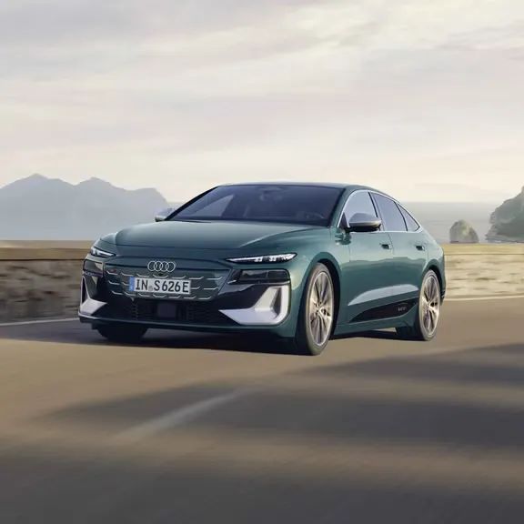 Audi S6 Sportback e-tron počas dynamickej jazdy po ceste popri pobreží. V pozadí je vidieť záliv a kopce.