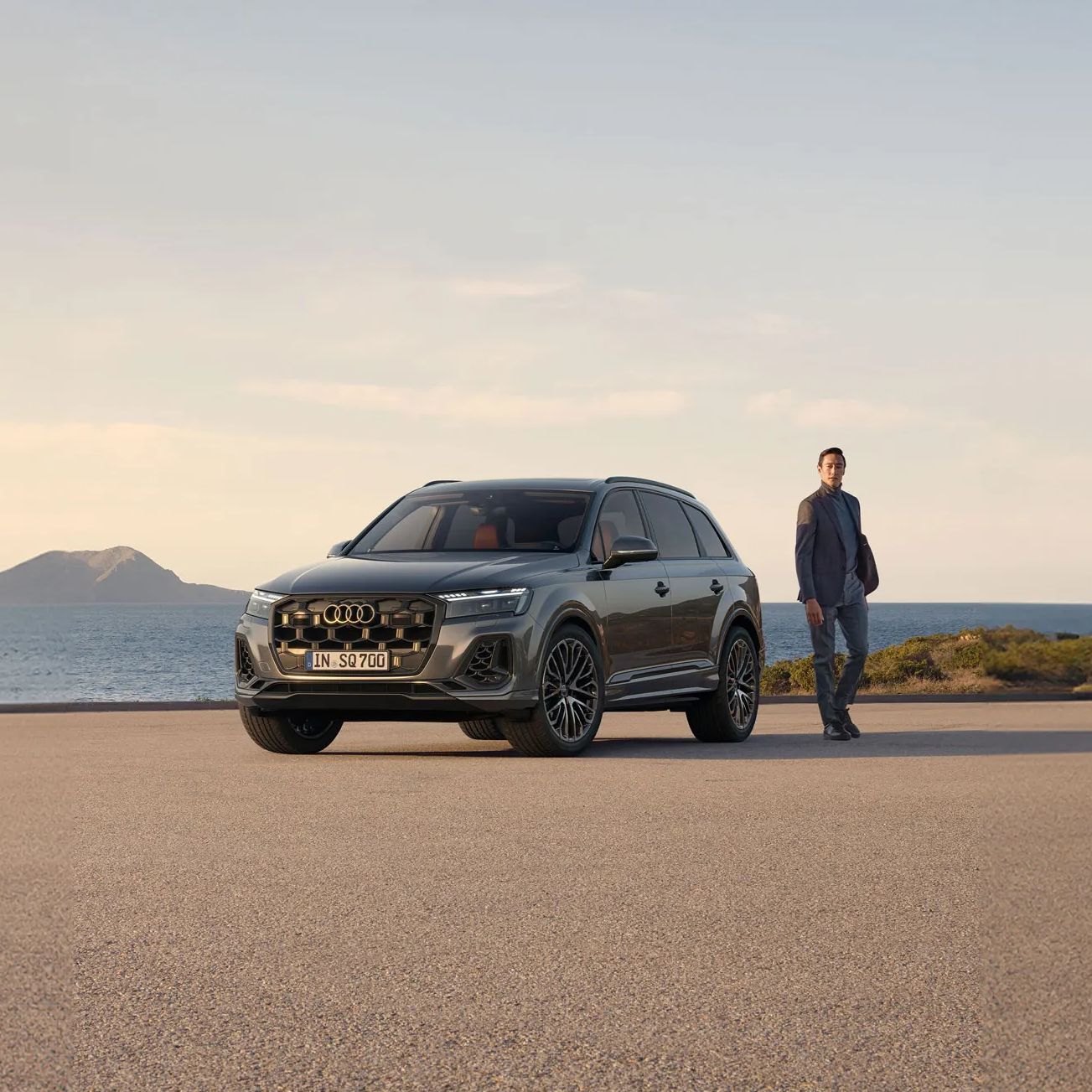 Audi SQ7 SUV zaparkované na voľnom priestore pri pobreží. Okolo vozidla prechádza muž. V pozadí je vidieť kopce a záliv.