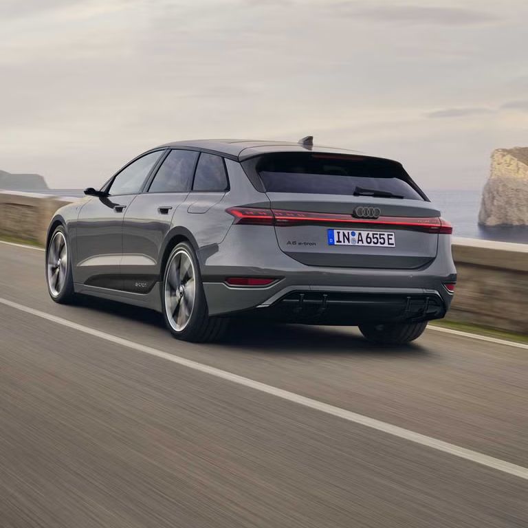 Schräge Heckaufnahme des A6 Avant e-tron, welcher auf einer Straße entlang des Meeres fährt. Im Hintergrund sind Felsen und Berge zu erkennen. 