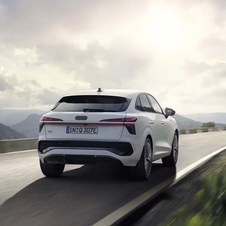 Audi Q3 Sportback e-hybrid počas jazdy po ceste. V pozadí je vidieť kopce a zatiahnuté oblohu, cez ktorú preniká slnečné svetlo a osvetľuje vozidlo. 