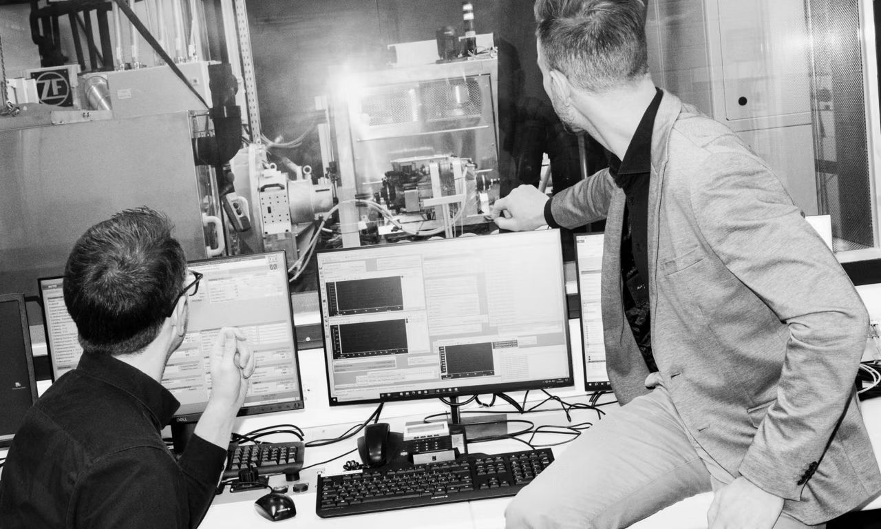 Dvaja muži sedia pred monitormi a cez okno sa pozerajú na testovaný systém v laboratóriu.