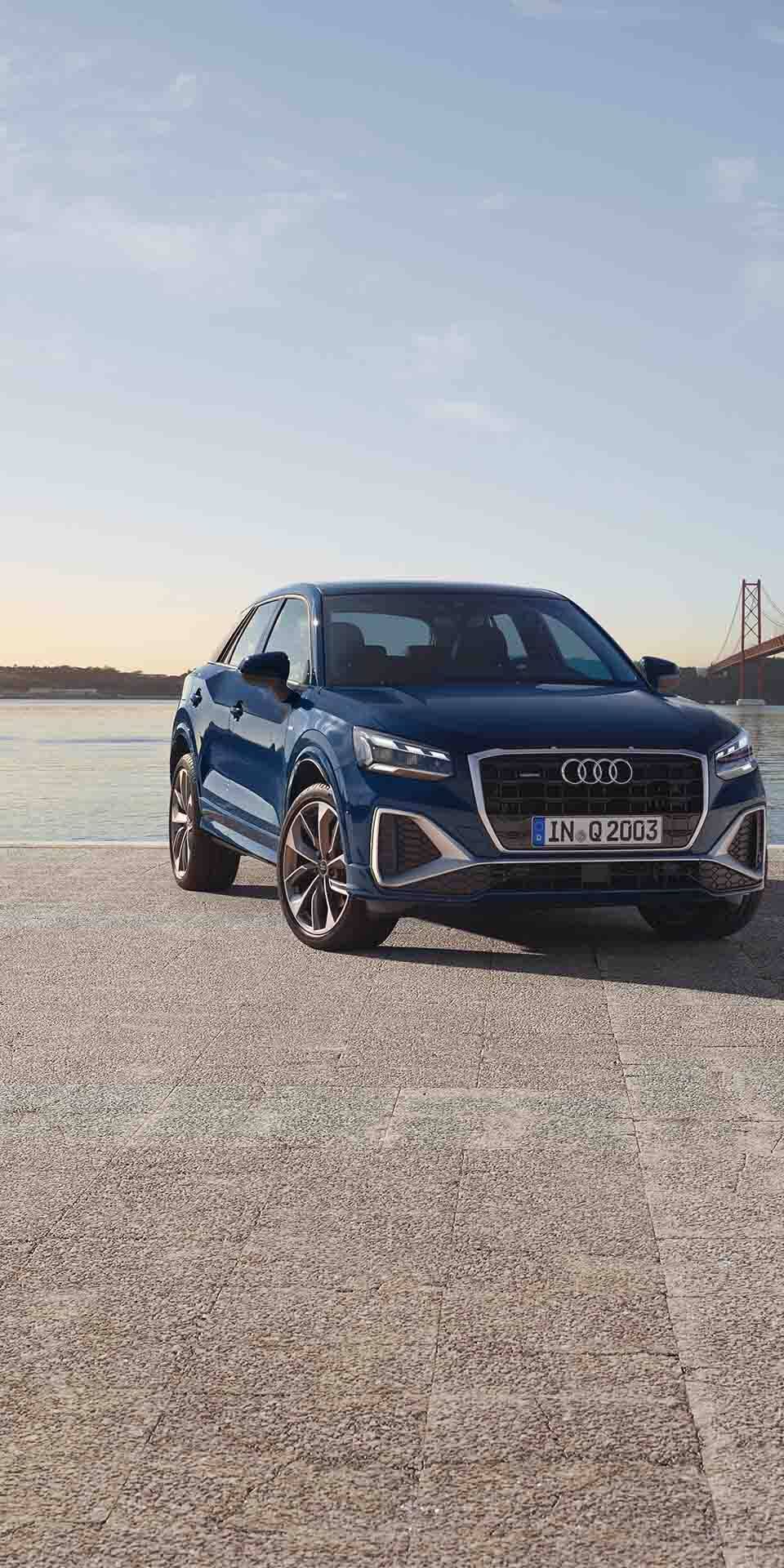 Modré Q2 značky Audi stoji na voľnej dláždenej ploche pri rieke a mostom cez ňu v pozadí. 