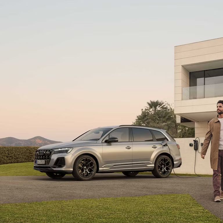 Bočný pohľad na strieborné Audi Q7 SUV TFSI e pri nabíjaní pred modernou vilou. 