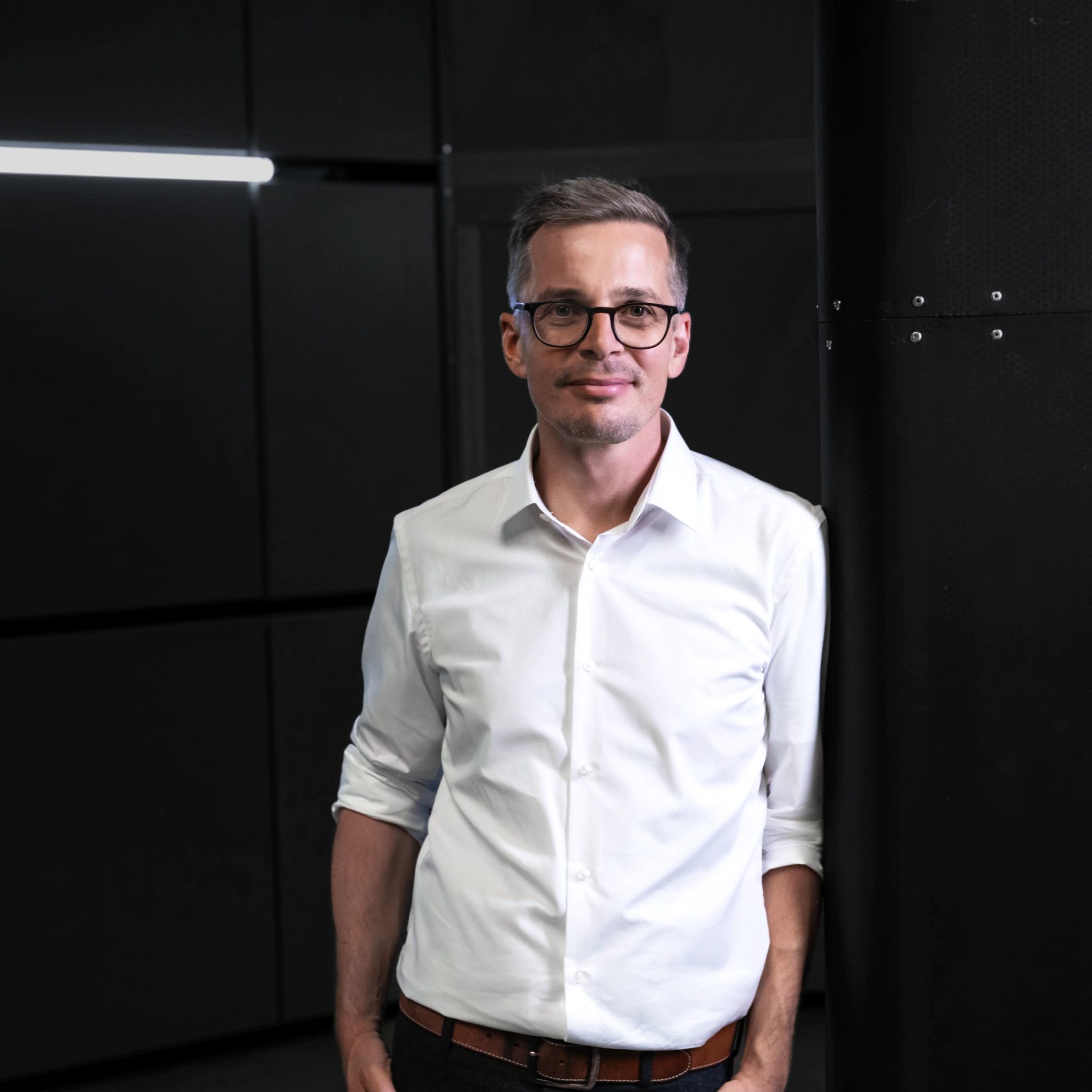 Muž v bielej košeli stojí opretý o stĺp v tmavej miestnosti. Pozadie je minimalistické a čierne.