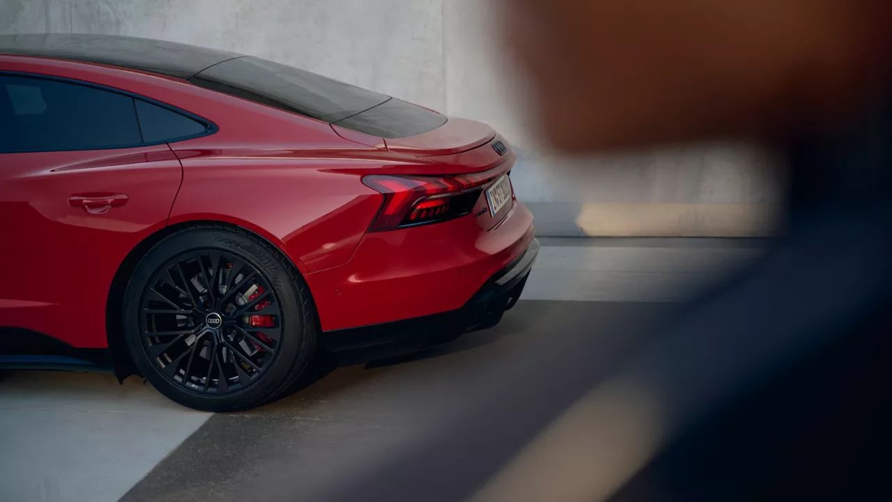Červené športové auto zaparkované pri stene. Detail na zadnú časť vozidla, viditeľné čierne disky kolies. V popredí rozmazaný objekt, pravdepodobne osoba.