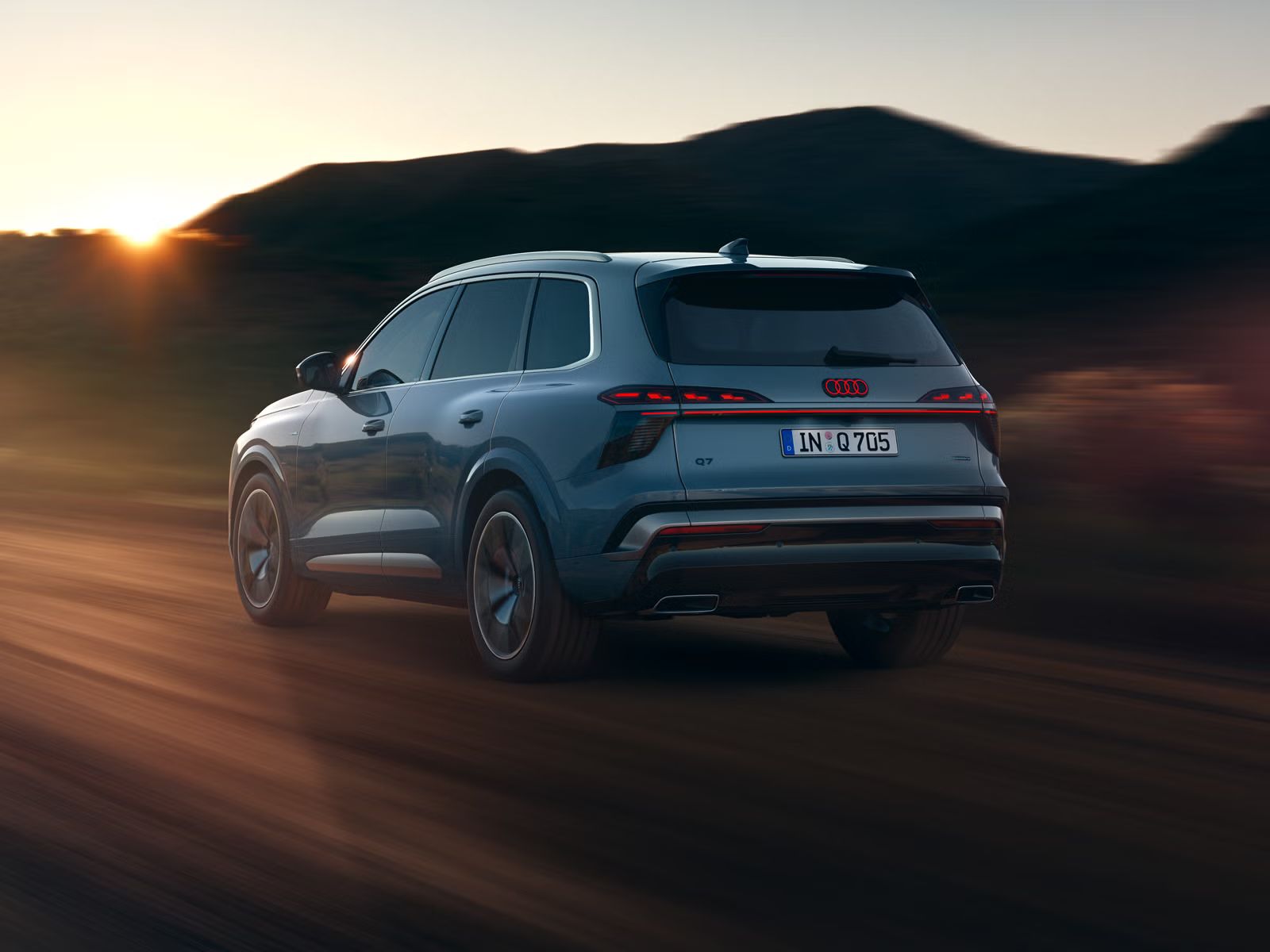 Audi Q7 SUV zachytený z boku zo zadnej strany pri plnej rýchlosti na vidieckej ceste, v pozadí sú hory. Scénu osvetľuje slnko zapadajúce za obzorom.  