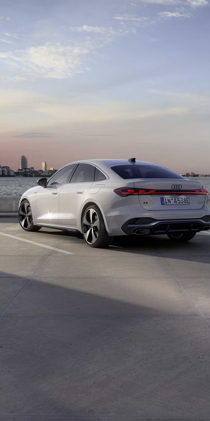 Die A5 Limousine e-hybrid steht im schrägen Heckprofil auf einem Parkplatz am Ufer eines Sees. Dahinter ist die Skyline einer Stadt bei Dämmerung zu sehen.