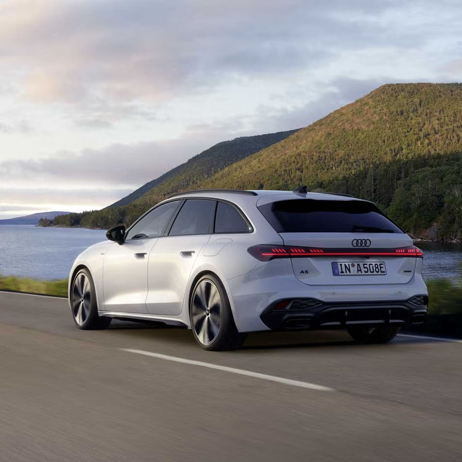 Audi A5 Avant e-hybrid počas jazdy na ceste popri pobreží. V pozadí vidieť záliv a zalesnené kopce.